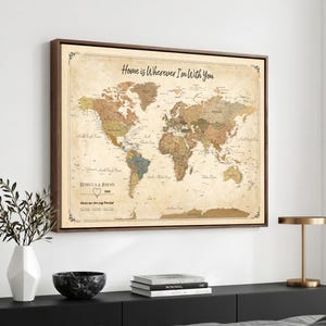 May include: A rectangular framed world map wall art with a vintage beige background. The map features muted earth tones, the text "Home is Wherever I'm With You" at the top, and "Rebecca & Jeremy" with a heart icon in the lower left corner.