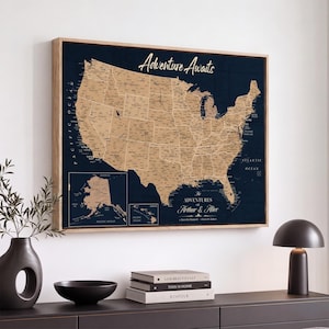 May include: A framed wall map of the United States in a tan color against a dark navy background. The map features the text Adventure Awaits at the top and The Adventures of Arthur & Alice at the bottom, with insets for Alaska and Hawaii.