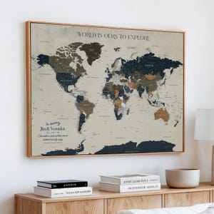 May include: A framed world map with a neutral color palette, featuring countries in shades of brown, blue, and gray. The text "WORLD IS OURS TO EXPLORE" is at the top. Below, the text reads "The Adventures of Jim & Veronica". The map is displayed above a wooden cabinet.