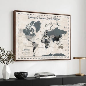 May include: A rectangular wall map in a dark wood frame featuring a world map with shades of gray and black landmasses on a cream background. The text Home is Wherever I'm With You is at the top, and The Adventures of the Jones Family is below.