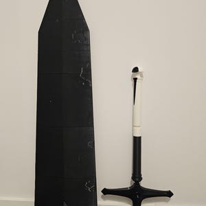 May include: A black, geometric sword and shield set. The shield is a tall, rectangular shape with a pointed top. The sword has a white handle and a black blade. The set is displayed against a white wall.