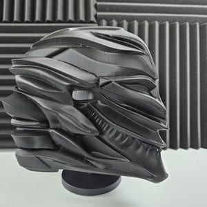 May include: A black helmet with a futuristic design, featuring layered, curved plates and ridges. The helmet has a textured appearance and is displayed on a small, round black stand. The design suggests a sci-fi or fantasy theme.