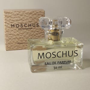 Könnte beinhalten: Ein klarer, rechteckiger Parfümflakon mit einer facettierten, klaren Kappe und einem goldfarbenen Zerstäuber. Die Flasche ist mit "MOSCHUS EAU DE PARFUM" beschriftet und enthält 50 ml Parfüm. Eine strukturierte, beige Schachtel mit dem gleichen Branding steht dahinter.