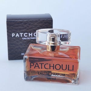 Könnte beinhalten: Ein orangefarbener Glasparfümflakon mit einem transparenten Deckel und goldenen Akzenten. Die Flasche ist mit "PATCHOULI EAU DE PARFUM" beschriftet und enthält 50 ml Duft. Eine strukturierte dunkelbraune Schachtel befindet sich hinter der Flasche.