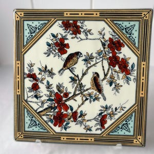 May include: A square ceramic tile featuring a cream-colored center with a detailed illustration of two brown birds perched on branches with red flowers and green leaves. The tile is framed by a geometric border in tan, black, and light blue.