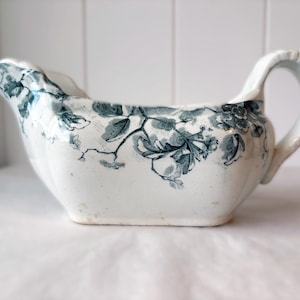 May include: A white ceramic gravy boat featuring a dark green floral and fruit pattern. The vessel has a curved handle, a wide pouring spout, and a scalloped rim. The exterior surface is decorated with detailed botanical illustrations.