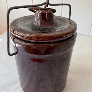 May include: A dark brown glazed ceramic crock with a matching lid and a metal wire bail handle. The cylindrical container features a smooth, glossy finish and a central knob on the lid, with a weathered, oxidized appearance on the metal handle.