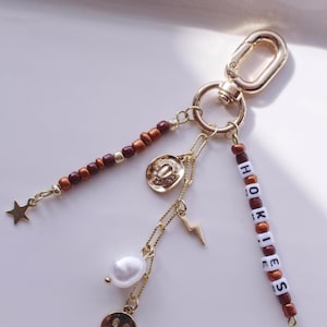 May include: Gold-toned keychain with multiple charms. Charms include a star, a cowboy hat, a lightning bolt, a smiley face, and a pearl. The keychain also has beads and the word "HOKIES" spelled out in white and brown beads.