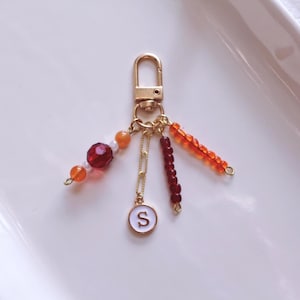 May include: A gold-colored keychain with a lobster clasp. The keychain features a chain with a small, white-rimmed charm with the letter "S". It also has strands of red and orange beads.