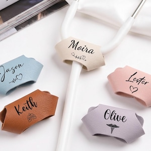 Puede incluir: Cinco etiquetas de identificación para estetoscopio de piel sintética rectangular en azul claro, tostado, beige, rosa y lavanda. Cada etiqueta presenta un nombre personalizado en letra cursiva negra y un pequeño icono médico.