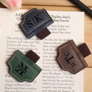 May include: Three leather bookmarks in navy, green, and brown, each with a personalized initial and name engraving. The bookmarks have a stitched border and an elastic band for securing to a book.