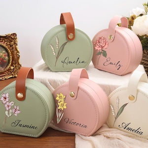 Könnte beinhalten: Fünf runde Schmuckkästchen in Pastellfarben mit floralen Designs und Namen. Jedes Kästchen hat einen Griff und einen goldenen Verschluss. Die Kästchen sind in Grün-, Rosa- und Weißtönen gehalten und tragen Namen wie "Amelia" und "Jasmine".