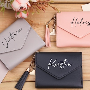 Peut inclure: Trois petits portefeuilles personnalisés, gris, rose et noir, chacun avec un nom en écriture cursive. Les portefeuilles ont une fermeture à rabat et un pompon avec un charme en forme de cœur. Sur une surface en bois.