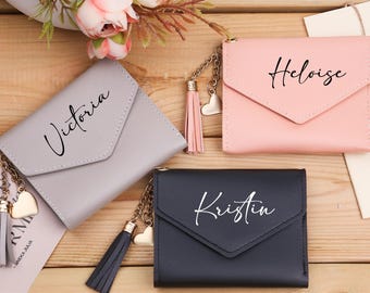 May include: Three small, personalized wallets in gray, pink, and black, each with a name in cursive script. The wallets have a flap closure and a tassel with a heart charm. The wallets are on a wooden surface.