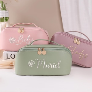 以下が含まれることがあります： ピンク、セージグリーン、ラベンダーの3つのパーソナライズされた化粧ポーチ。各バッグにはハンドル、ゴールドのジッパー、筆記体で名前が書かれています。ピンクのバッグには「Betsy」、グリーンのバッグには「Muriel」、ラベンダーのバッグには「Gladys」と書かれています。