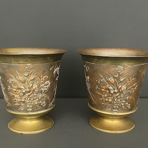Peut inclure: Une paire de jardinières métalliques assorties avec un bord large et évasé et une base circulaire sur pied. La surface extérieure présente un motif floral en relief avec des fleurs et des feuillages cuivrés sur un fond en laiton vieilli.