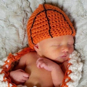 May include: A newborn baby wearing a crocheted orange and brown basketball hat. The baby is swaddled in a white and orange crocheted blanket.
