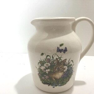 May include: Cream-colored ceramic pitcher with a curved handle and a spout. The pitcher features a colorful illustration of a brown bunny surrounded by flowers and butterflies. A decorative item for the kitchen or dining room.