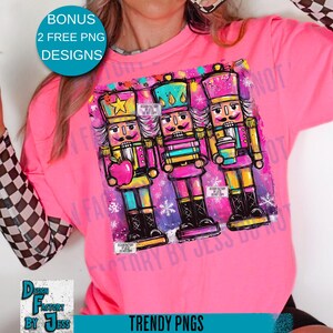 May include: A bright pink long-sleeve shirt featuring a colorful graphic of three nutcrackers. The nutcrackers are in shades of pink, yellow, blue, and green. The shirt has the text "TRENDY PNGS" and "BONUS 2 FREE PNG DESIGNS".