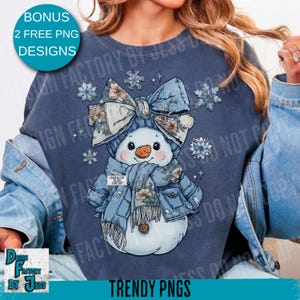 May include: A blue sweatshirt featuring a snowman design. The snowman wears a blue hat, scarf, and jacket, all adorned with floral patterns. The image includes text that says "BONUS 2 FREE PNG DESIGNS" and "TRENDY PNGS".