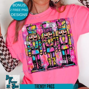 May include: A pink t-shirt featuring a colorful graphic of three nutcrackers with festive lights and snowflake accents. The design includes the text "DESIGN FACTORY BY JESS DO NOT COPY" and "TRENDY PNGS".