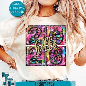 May include: A cream-colored t-shirt featuring a colorful graphic design. The design includes the numbers "2026" in leopard print, the word "hello" in gold glitter, and a decorative ornament. The shirt also has text that says "BONUS 2 FREE PNG DESIGNS".