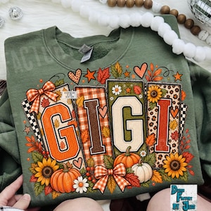 May include: An olive green crewneck sweatshirt featuring a graphic print of the word GIGI in large block letters. The design is decorated with autumn-themed elements including orange pumpkins, sunflowers, plaid patterns, leopard print, and orange bows.