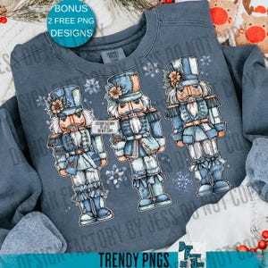 May include: A blue-grey sweatshirt featuring three illustrated nutcrackers in blue and white uniforms. Each nutcracker has a hat, a mustache, and a floral accent. The text "DESIGN FACTORY BY JESS DO NOT COPY" is visible. The sweatshirt also has the text "TRENDY PNGS".