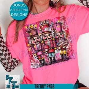May include: A bright pink long-sleeved shirt featuring a colorful graphic of three nutcracker figures. The nutcrackers are dressed in red, pink, and yellow uniforms. The shirt also has the text "BONUS 2 FREE PNG DESIGNS" and "TRENDY PNGS".