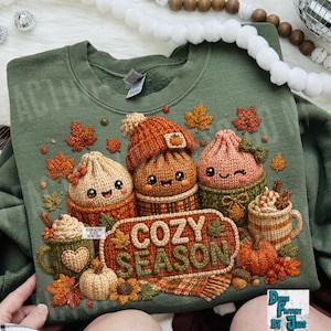 May include: An olive green crewneck sweatshirt featuring a graphic print of three smiling, crocheted-style characters in mugs, a mug of hot cocoa with whipped cream, pumpkins, and autumn leaves. The text COZY SEASON is centered in a textured frame.