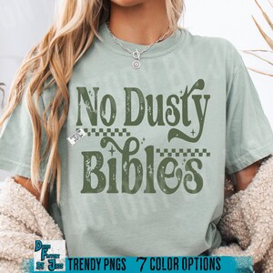 May include: A light green t-shirt with the text "No Dusty Bibles" in a retro font. The design includes decorative elements and is printed in a darker green color. The shirt has a relaxed fit.
