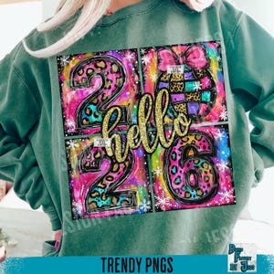 May include: A teal sweatshirt with a colorful graphic design. The design features the numbers "2026" with leopard print accents, a decorative ornament, and the word "hello" in gold glitter. The design is set against a vibrant, multi-colored background.