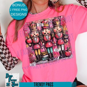 May include: A bright pink long-sleeve shirt featuring a colorful graphic of three nutcracker figures. The nutcrackers are in red, gold, and black, with apples and books. The shirt also has the text "TRENDY PNGS" and "BONUS 2 FREE PNG DESIGNS".
