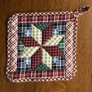 May include: Square quilted pot holder with a starburst design. The center features a patchwork star in shades of red, green, blue, and beige plaid fabrics. The border is a red and white gingham pattern, with a loop for hanging.