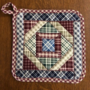 May include: A square, quilted potholder with a loop for hanging. The design features a central square of blue plaid surrounded by concentric squares and diamonds of various plaid patterns in navy, burgundy, green, and tan. The edges are trimmed with a burgundy and white gingham.