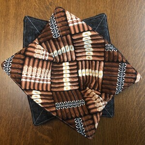 May include: A brown and black fabric coaster with a pattern of brown and white cigars. The coaster is made of a soft, quilted fabric and has a square shape.