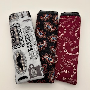 May include: Three rectangular fabric cases with dark top edges. The left case is light gray with black barber shop motifs, including mustaches and text. The middle case features a black background with a colorful paisley pattern. The right case is maroon.