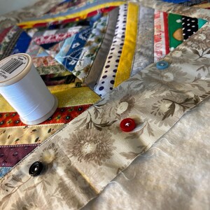 May include: A multicolored fabric quilt featuring various patterned strips and a beige floral border. A spool of white hand quilting thread sits on the quilt. Three buttons in red, blue, and black are attached to the floral fabric border.
