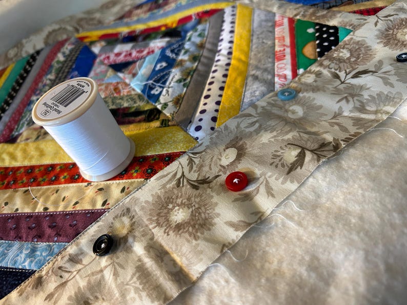May include: A multicolored fabric quilt featuring various patterned strips and a beige floral border. A spool of white hand quilting thread sits on the quilt. Three buttons in red, blue, and black are attached to the floral fabric border.