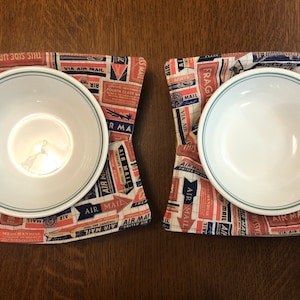 May include: Two white bowls with a green rim are placed on top of two fabric placemats. The placemats are made of a cream-colored fabric with a red, blue, and white pattern of vintage airmail labels. The text on the labels includes "Air Mail", "Via Air Mail", and "This Side Up".