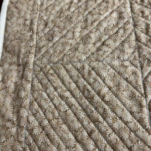 May include: A quilted fabric featuring a repeating tan and cream leaf pattern. The material is stitched with a diagonal grid design, creating a textured surface. A solid cream-colored fabric border runs along the left edge of the textile.
