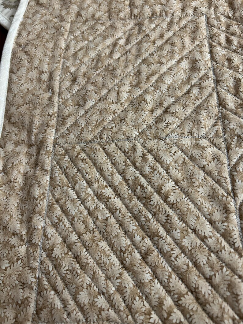 May include: A quilted fabric featuring a repeating tan and cream leaf pattern. The material is stitched with a diagonal grid design, creating a textured surface. A solid cream-colored fabric border runs along the left edge of the textile.