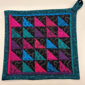 May include: A square quilted potholder with a teal border and hanging loop. The center features a patchwork design of black triangles with paisley patterns, alternating with triangles of pink, blue, teal, and purple.