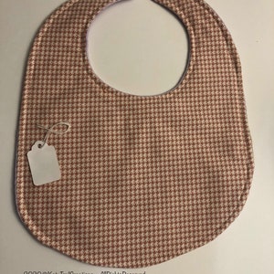May include: A pink and brown houndstooth bib with a white tag attached. The bib has two white snaps on the back.