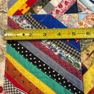 May include: A multicolored fabric quilt featuring diagonal strips of various patterns including polka dots, florals, and solid colors. A yellow measuring tape is placed across the quilt, showing a width measurement of approximately 6 inches.