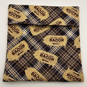 May include: A brown and black plaid sandwich bag with a fold-over closure. The bag is patterned with tan-colored pig-shaped graphics that read "FARM FRESH - SMOKED BACON". The bag is designed to hold food items.