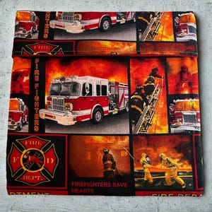 May include: A square, folded fabric item featuring a collage of images related to firefighting. The design includes red fire trucks, firefighters, flames, and the text "Fire Dept." and "Firefighters Save Hearts" on a black background.