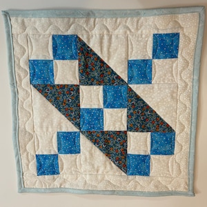 Puede incluir: Una pequeña pared acolchada con un patrón geométrico azul y blanco. La colcha está hecha de cuadrados y triángulos y tiene un borde azul claro.