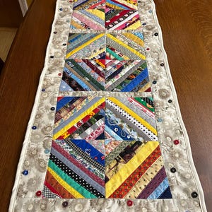 May include: A rectangular quilted table runner featuring three central square blocks made of multi-colored fabric strips in a diagonal pattern. The runner is bordered by a beige floral fabric and decorated with small, scattered buttons in various colors.