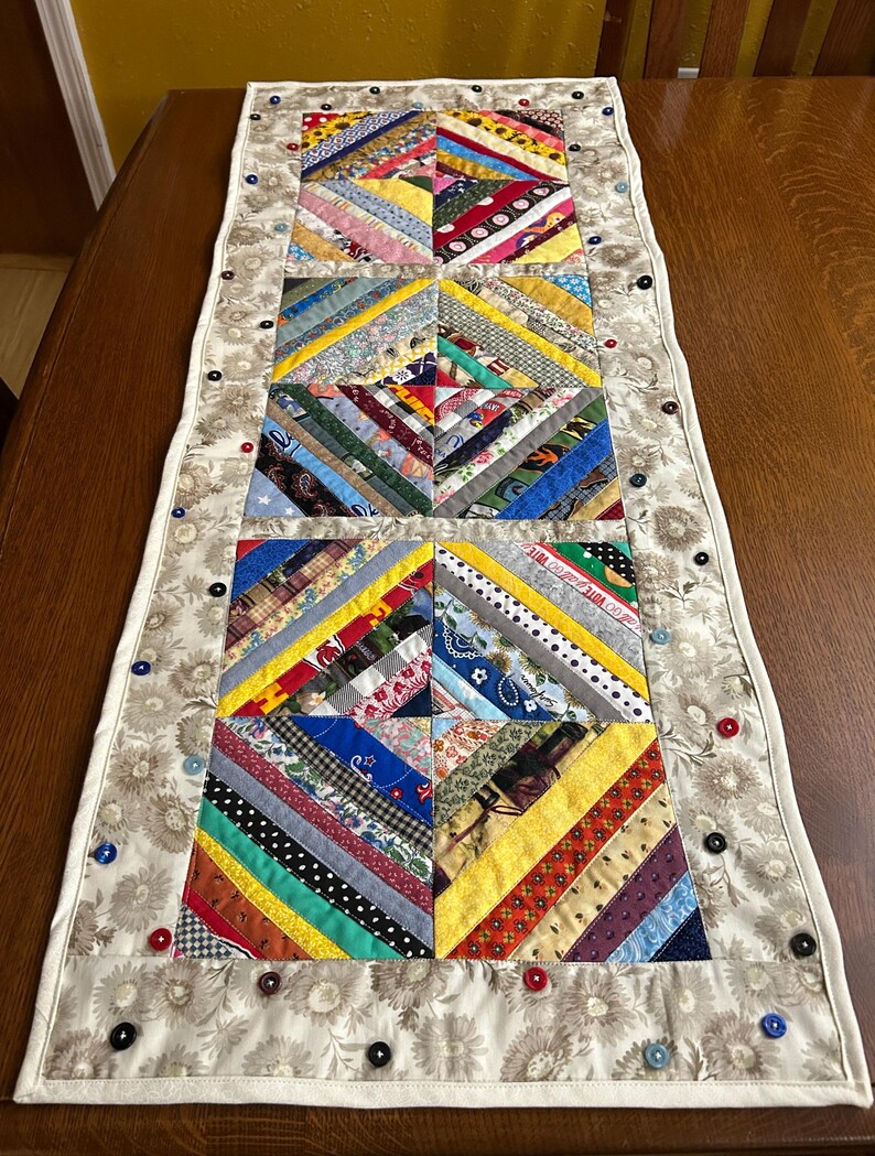 May include: A rectangular quilted table runner featuring three central square blocks made of multi-colored fabric strips in a diagonal pattern. The runner is bordered by a beige floral fabric and decorated with small, scattered buttons in various colors.